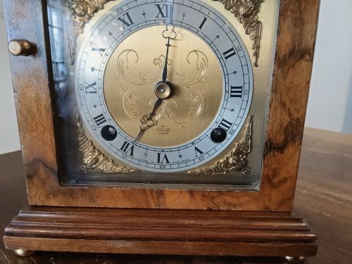 fig 1. A rectangular burr walnut bracket clock featuring a top brass handle, arched brass dial face with Roman numerals, and a silvered chapter ring.