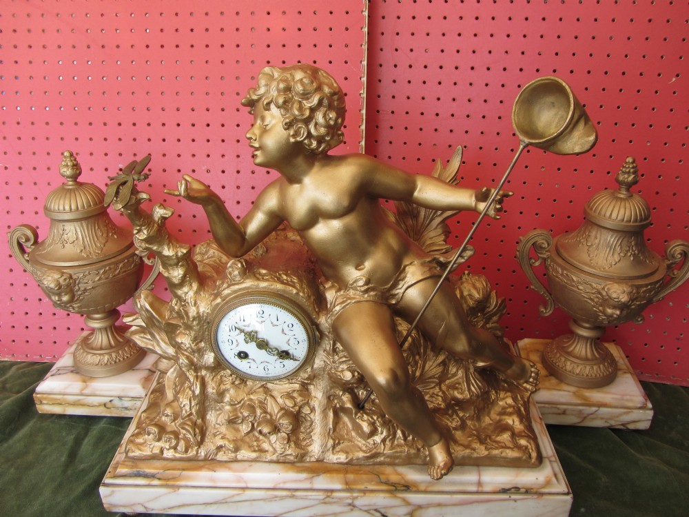 A gilt metal figural mantel clock depicting a cherub holding a net towards a dragonfly, set on a marble base and flanked by two matching gilt urns.