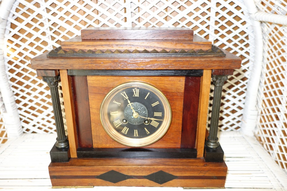 An antique French mantel clock with mixed specimen wood veneers, dark fluted side columns, a stepped pediment, and a dark circular dial framed in brass.