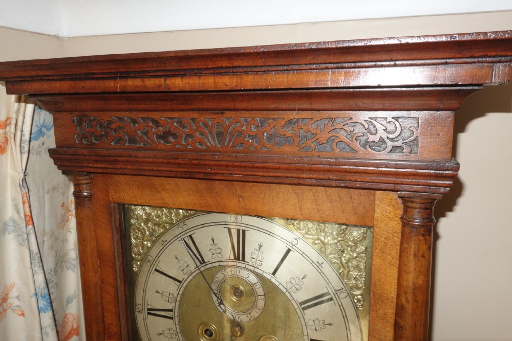 An early Georgian walnut longcase clock featuring a brass dial with Roman numerals, flanked by columns on the hood and a veneered wooden cabinet door. View X