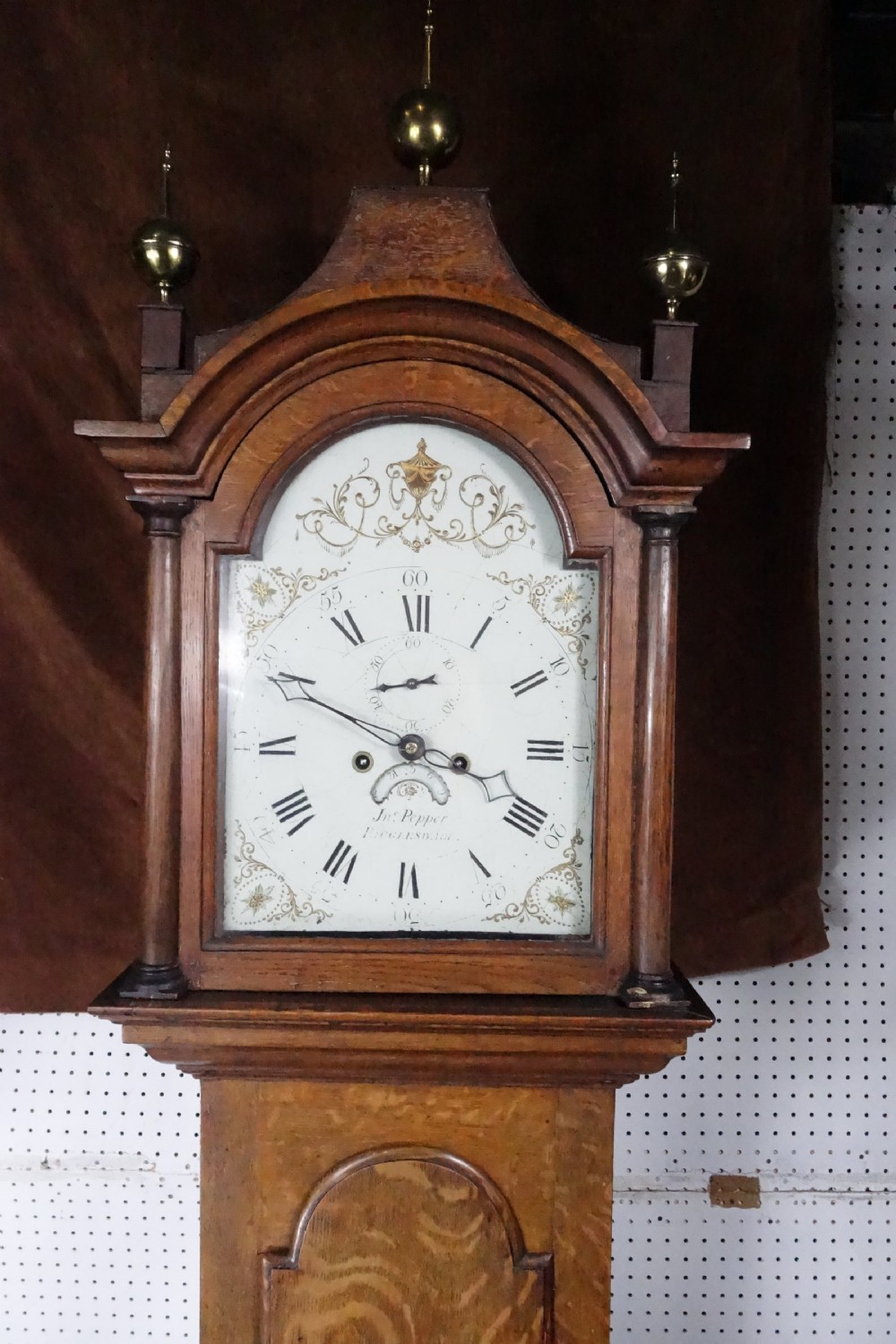 Georgian light oak longcase clock hood with brass finials, arched glass door, and white painted dial featuring Roman numerals.