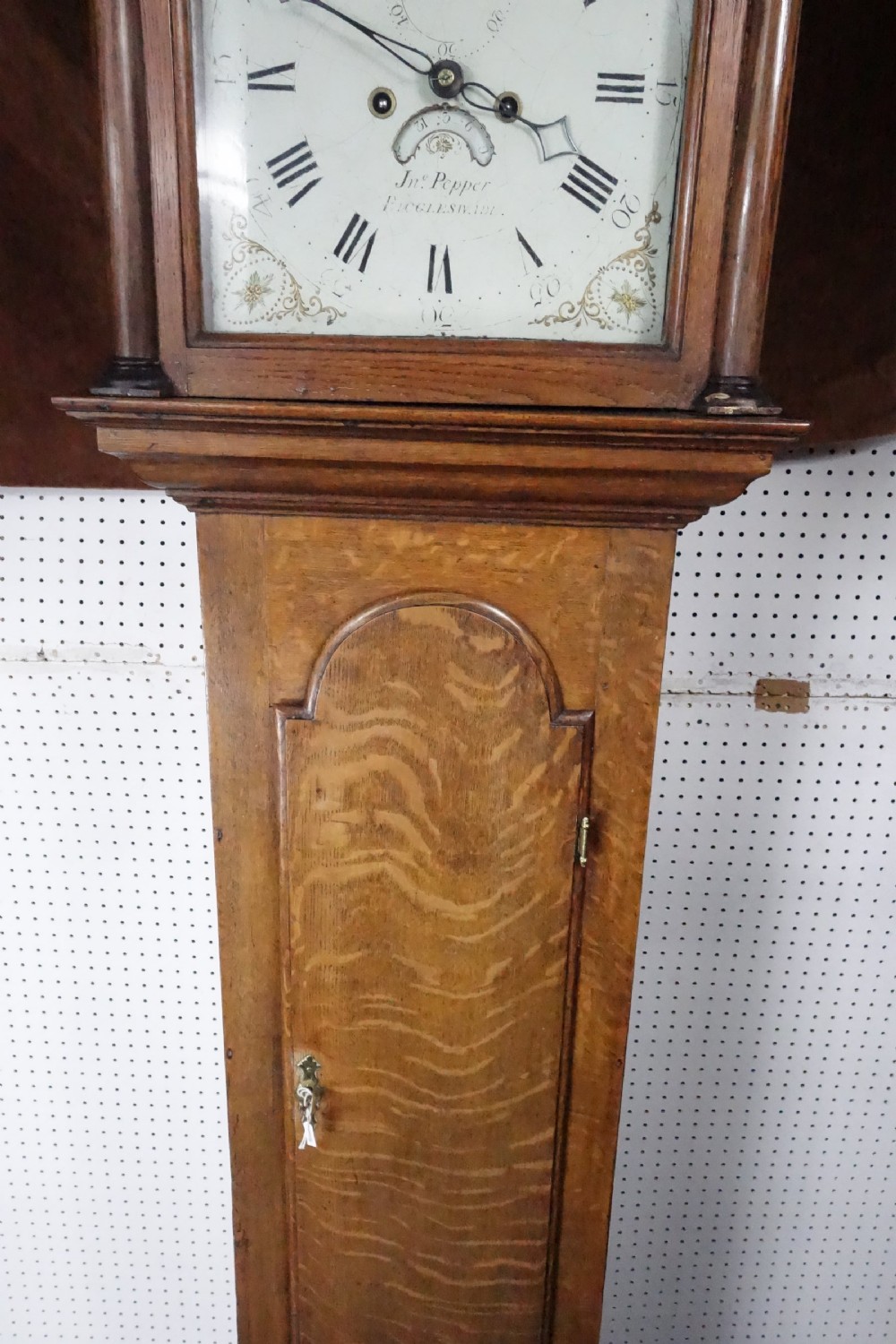 Georgian light oak longcase clock hood with brass finials, arched glass door, and white painted dial featuring Roman numerals. View V