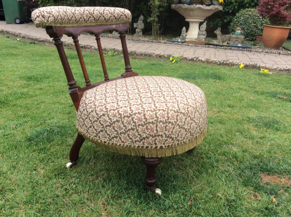 A Victorian low-seated nursing chair featuring a dark mahogany spindle back, padded top rail, pattern-upholstered seat with fringe trim, and turned front legs with castors.