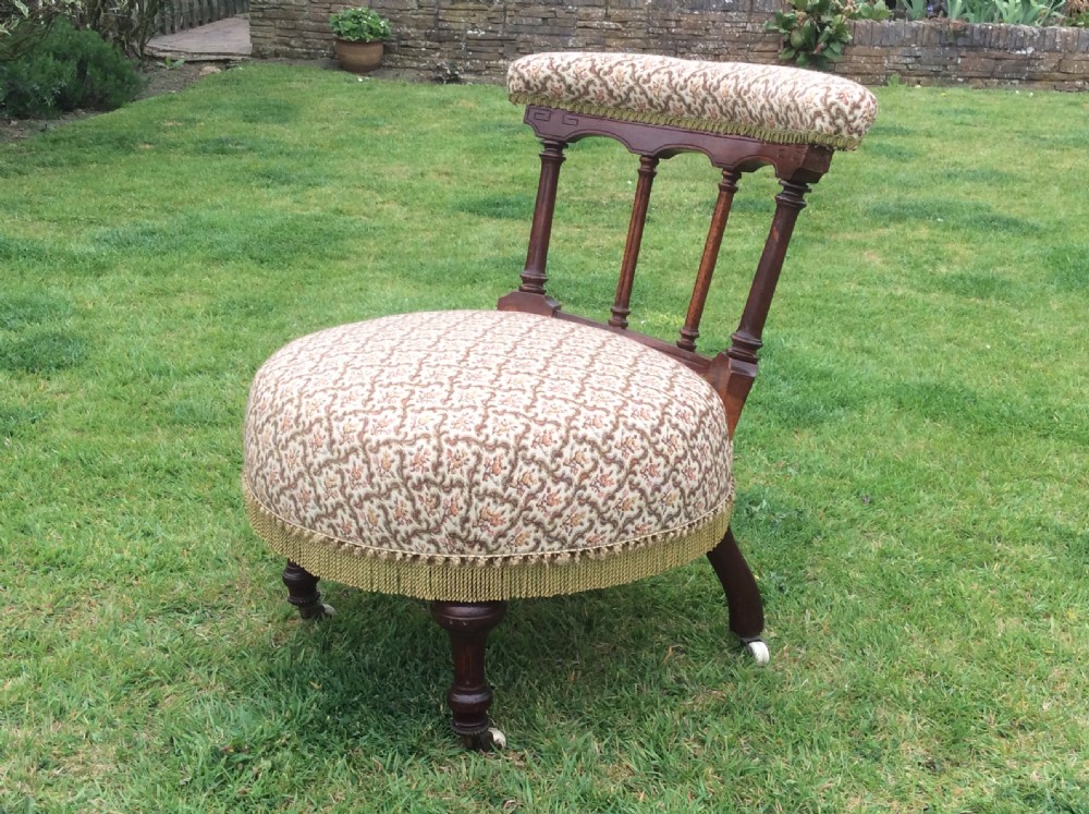 A Victorian low-seated nursing chair featuring a dark mahogany spindle back, padded top rail, pattern-upholstered seat with fringe trim, and turned front legs with castors. View VII