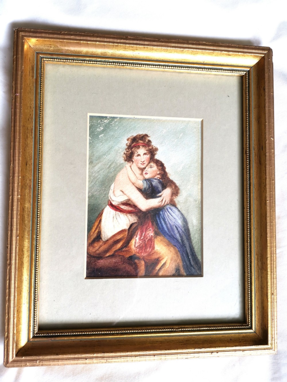 A small 19th-century watercolour portrait depicting a mother embracing her young child set within a wide cream mount and a beaded gold gilt frame.