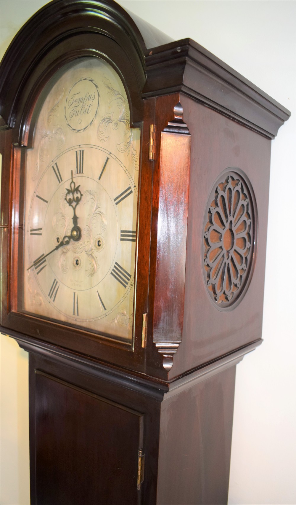 An antique dark mahogany longcase clock featuring an arched hood, engraved silvered dial with Roman numerals, and side fretwork sound apertures.