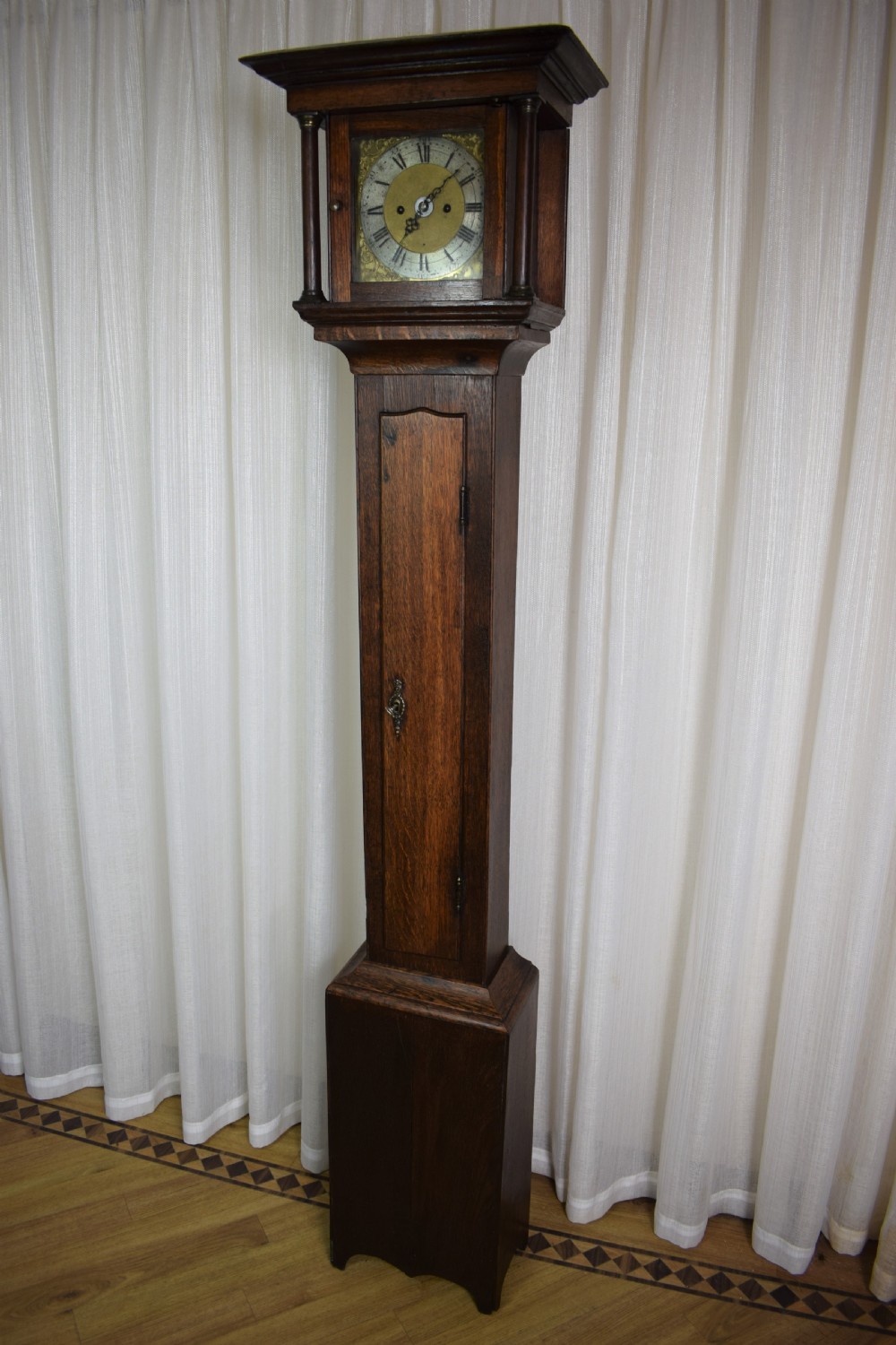 A dark brown timber grandmother clock featuring a square brass dial framed by glass and flanked by turned columns. View II
