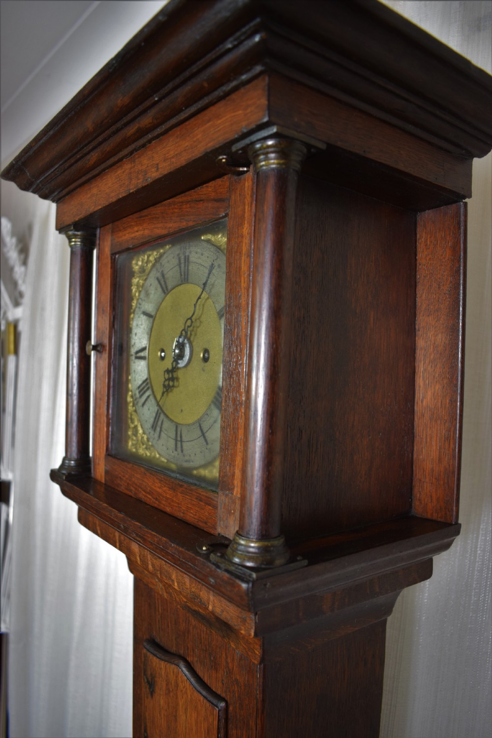 A dark brown timber grandmother clock featuring a square brass dial framed by glass and flanked by turned columns.