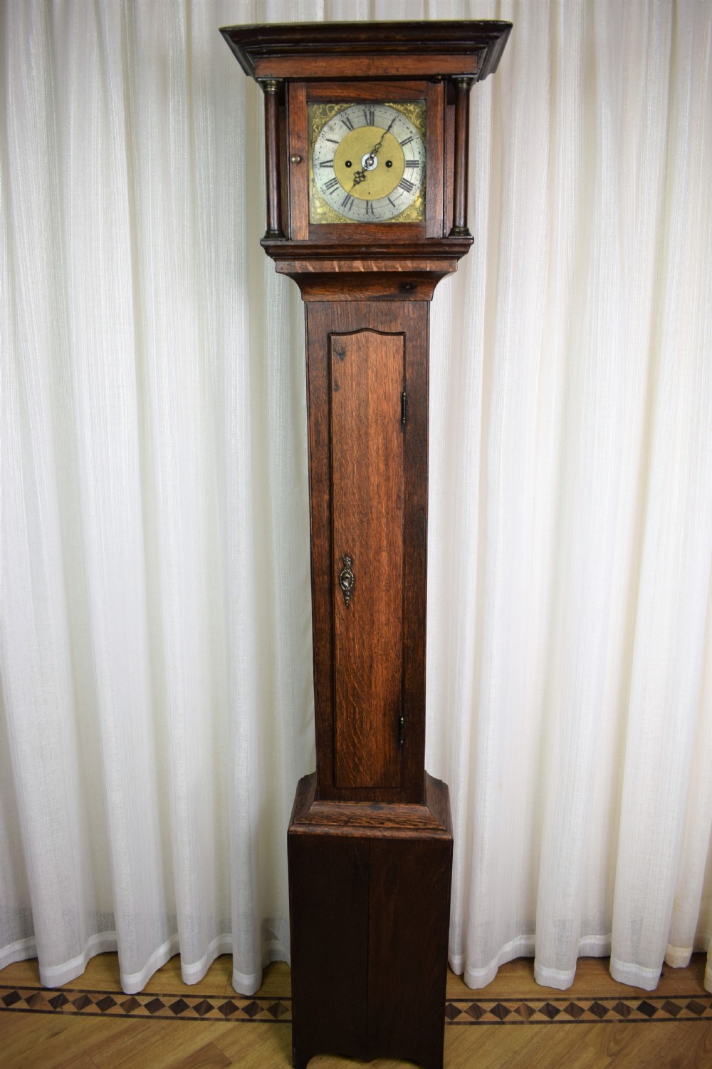 A dark brown timber grandmother clock featuring a square brass dial framed by glass and flanked by turned columns. View VI