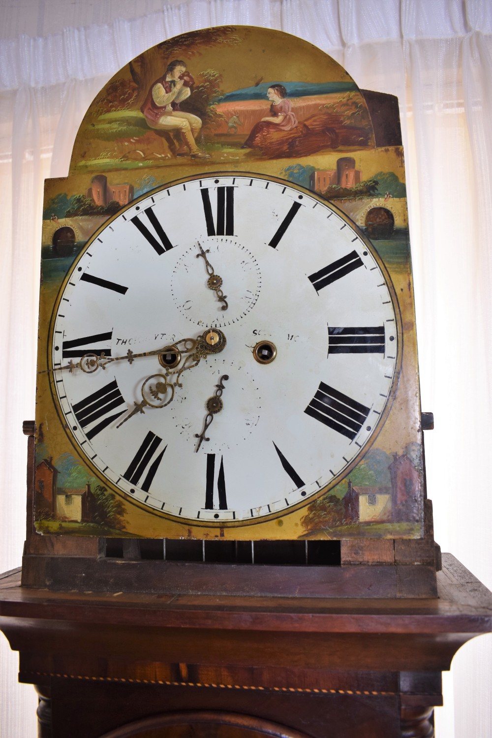 An early 19th-century Georgian flame mahogany longcase clock featuring a swan-neck pediment, brass patera, turned columns, and a hand-painted dial depicting a pastoral scene with Roman numerals. View XIV