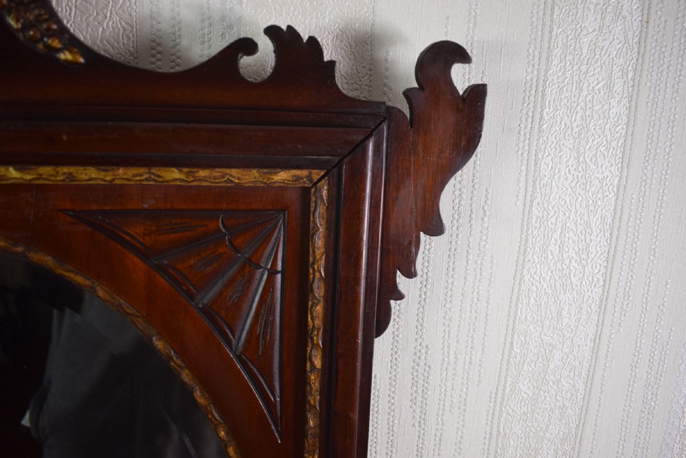 A 19th-century mahogany wall mirror featuring a bevelled oval glass plate, scroll fretwork border, gilded urn finial, and shell marquetry inlay. View VI