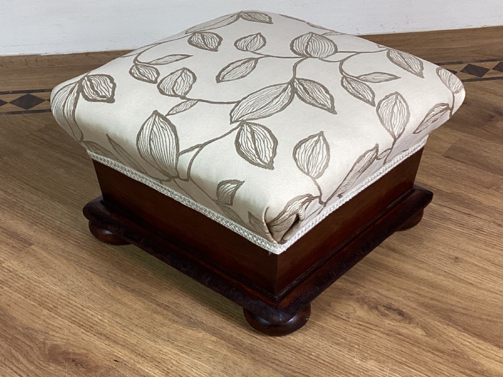 A square Victorian footstool featuring a dark mahogany plinth with turned bun feet and cream upholstery patterned with leaves.