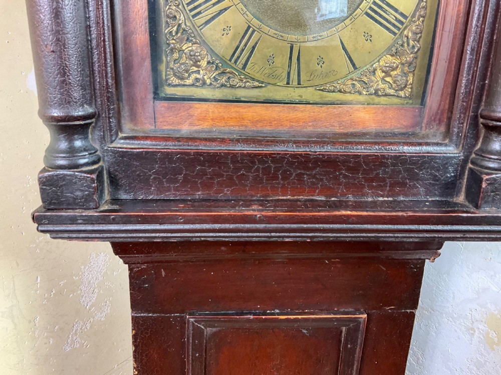 A tall dark brown painted pine longcase clock featuring a square brass dial with ornate corner spandrels, turned hood pillars, and a slender trunk door on a plinth base. View II
