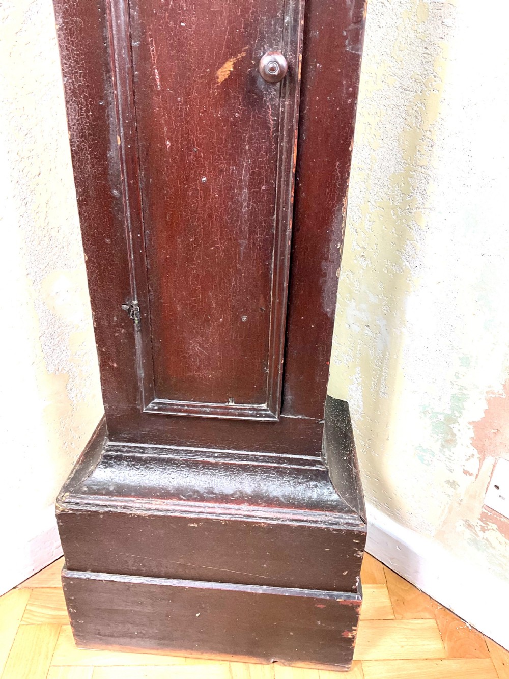 A tall dark brown painted pine longcase clock featuring a square brass dial with ornate corner spandrels, turned hood pillars, and a slender trunk door on a plinth base. View III