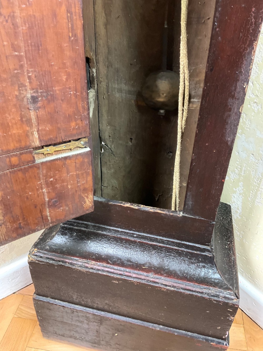 A tall dark brown painted pine longcase clock featuring a square brass dial with ornate corner spandrels, turned hood pillars, and a slender trunk door on a plinth base. View IV