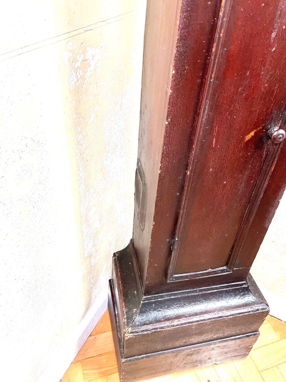 A tall dark brown painted pine longcase clock featuring a square brass dial with ornate corner spandrels, turned hood pillars, and a slender trunk door on a plinth base. View V