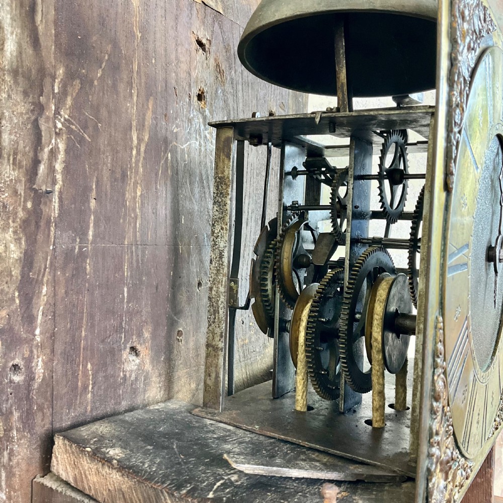 A tall dark brown painted pine longcase clock featuring a square brass dial with ornate corner spandrels, turned hood pillars, and a slender trunk door on a plinth base. View VIII