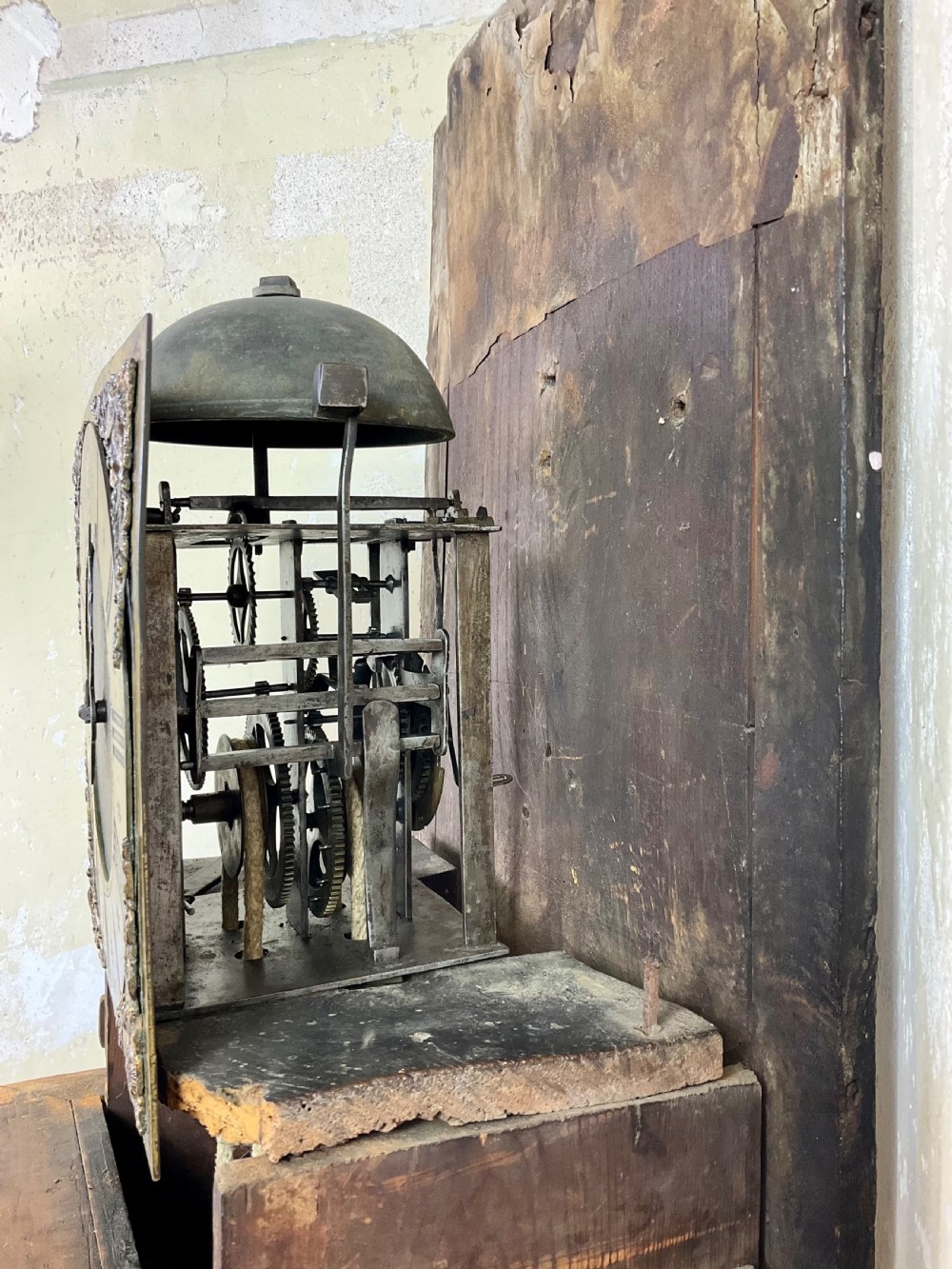 A tall dark brown painted pine longcase clock featuring a square brass dial with ornate corner spandrels, turned hood pillars, and a slender trunk door on a plinth base. View IX