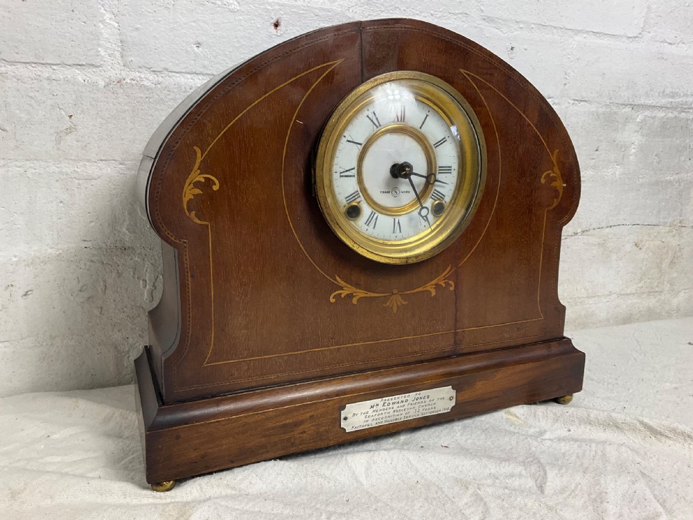 An Edwardian arched mahogany mantel clock featuring marquetry inlay, a circular brass-rimmed dial with Roman numerals, and a silver presentation plaque on the base.