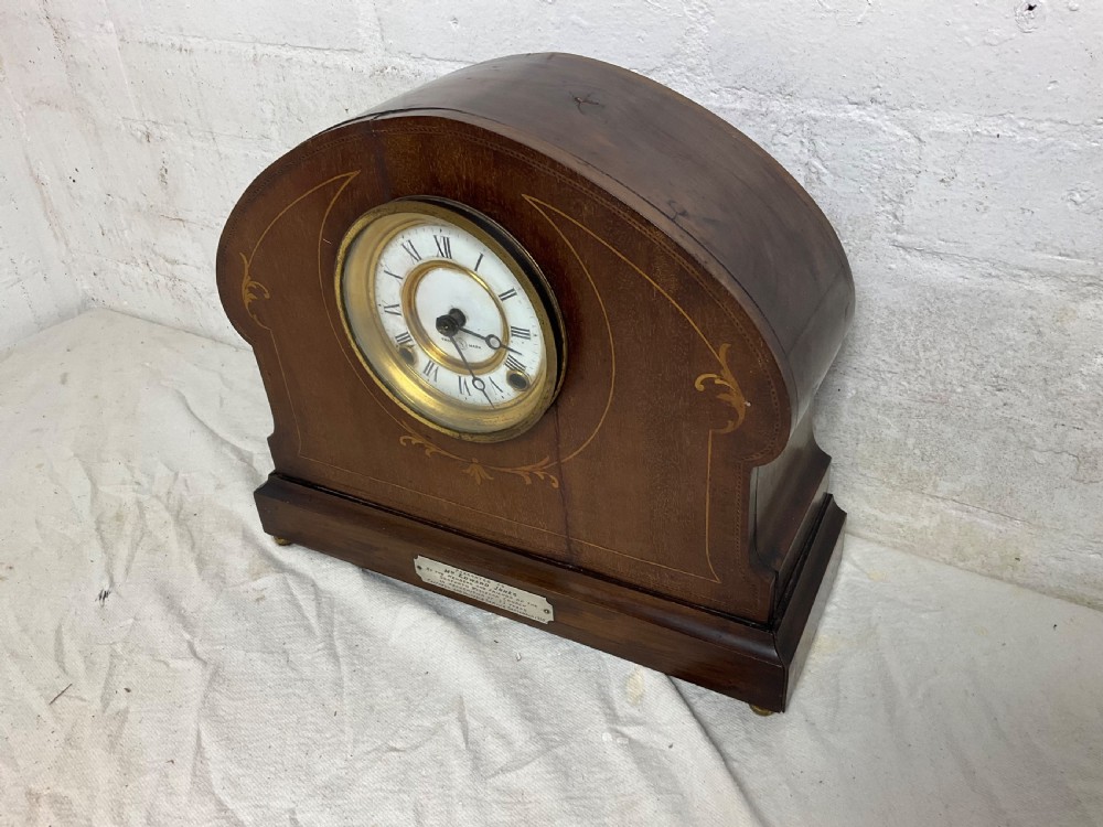 An Edwardian arched mahogany mantel clock featuring marquetry inlay, a circular brass-rimmed dial with Roman numerals, and a silver presentation plaque on the base. View VII
