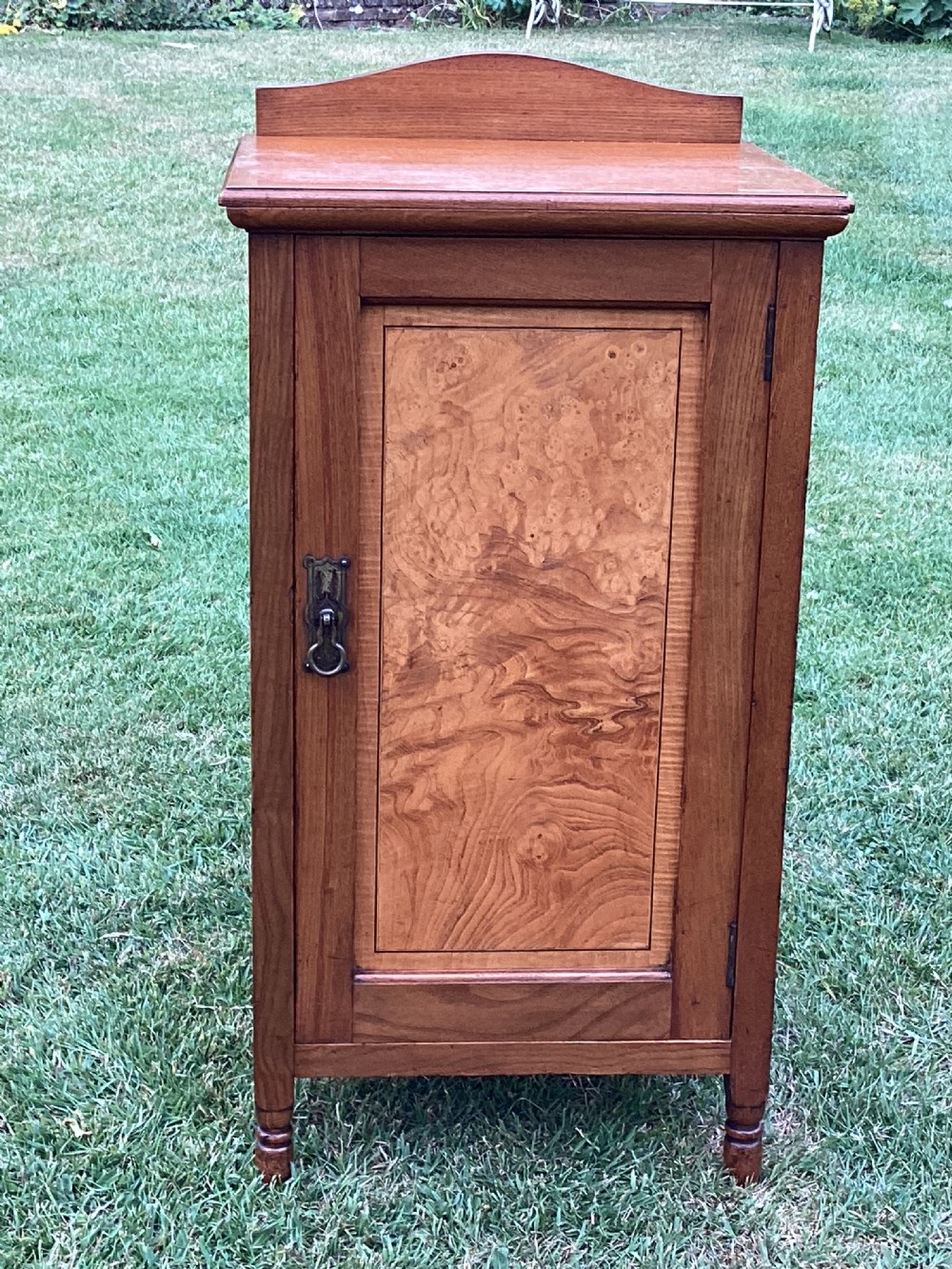 A light birch bedside cabinet featuring a single door with a central burr veneer panel, brass handle, and shaped upstand gallery.