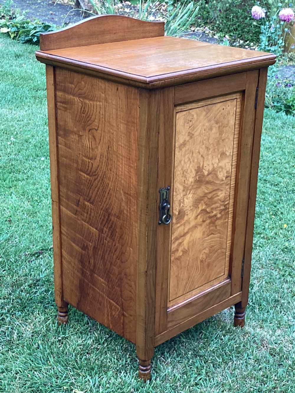 A light birch bedside cabinet featuring a single door with a central burr veneer panel, brass handle, and shaped upstand gallery. View II