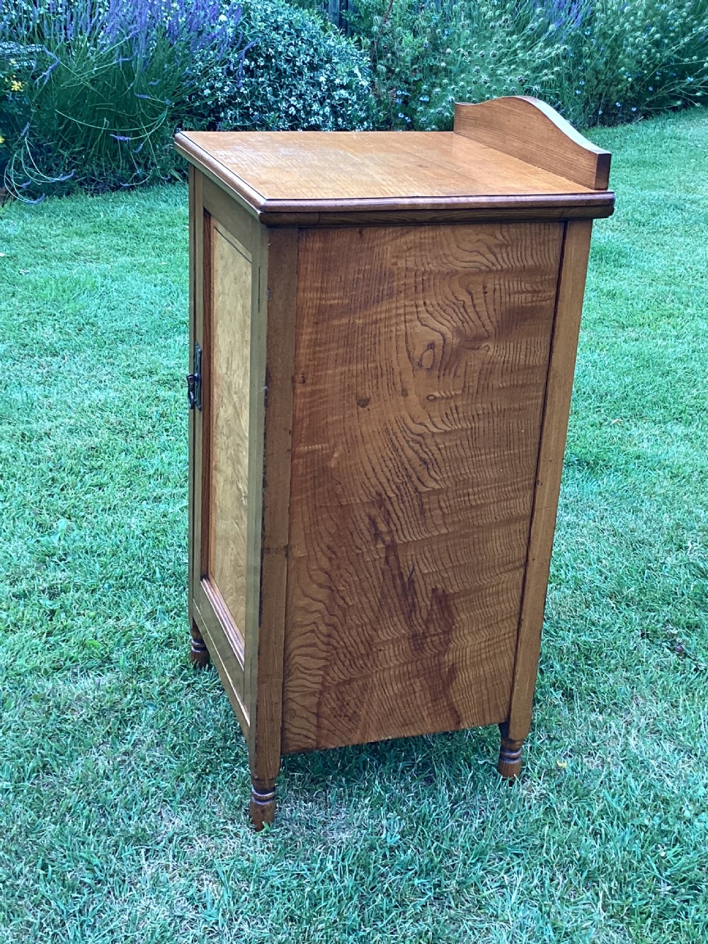 A light birch bedside cabinet featuring a single door with a central burr veneer panel, brass handle, and shaped upstand gallery. View III