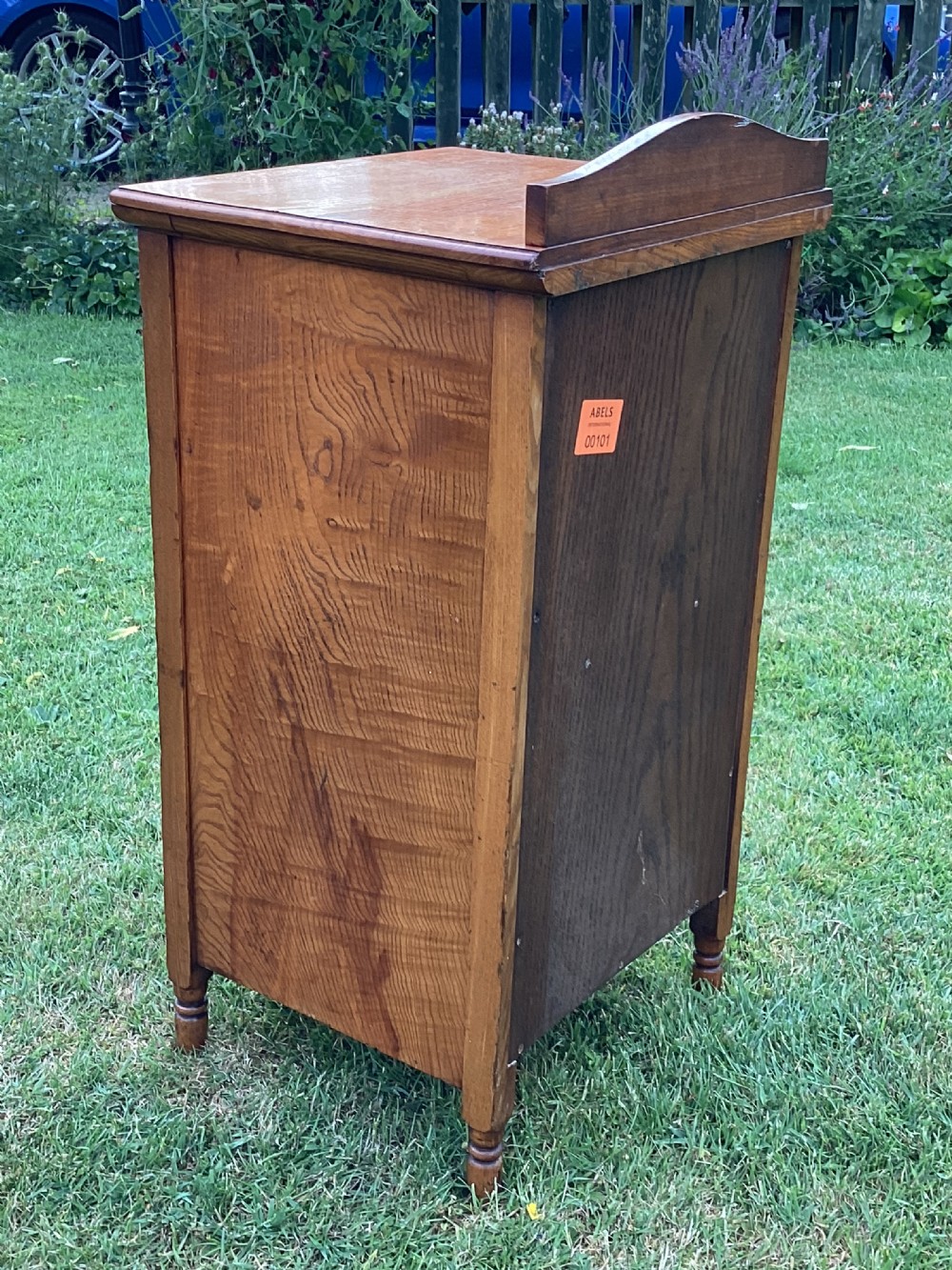 A light birch bedside cabinet featuring a single door with a central burr veneer panel, brass handle, and shaped upstand gallery. View IV