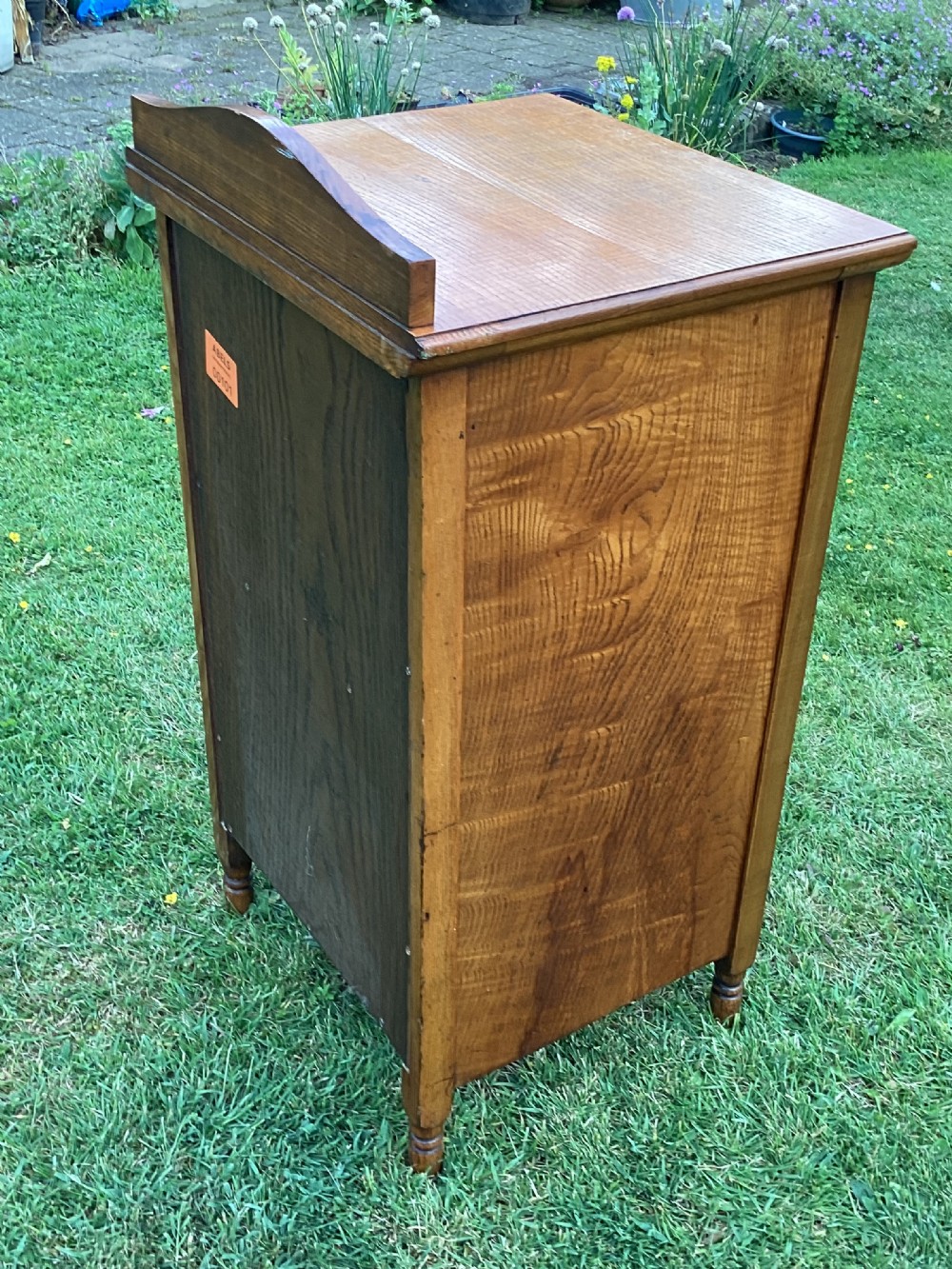 A light birch bedside cabinet featuring a single door with a central burr veneer panel, brass handle, and shaped upstand gallery. View V