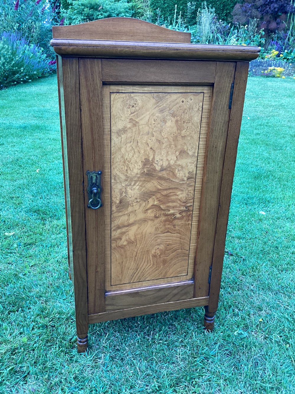 A light birch bedside cabinet featuring a single door with a central burr veneer panel, brass handle, and shaped upstand gallery. View X