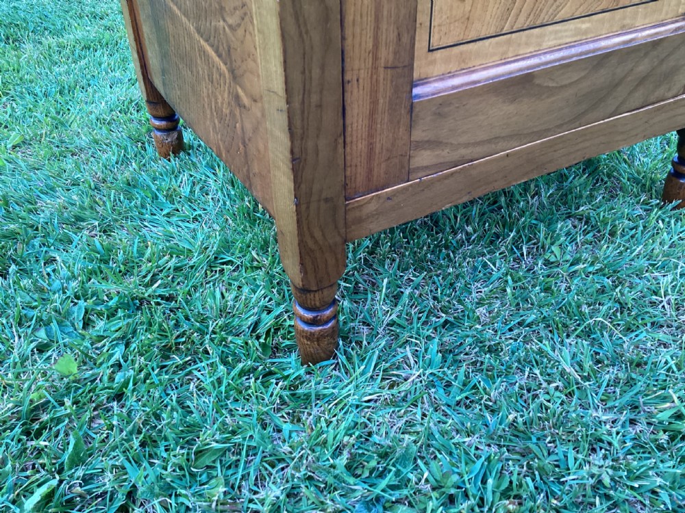 A light birch bedside cabinet featuring a single door with a central burr veneer panel, brass handle, and shaped upstand gallery. View XI