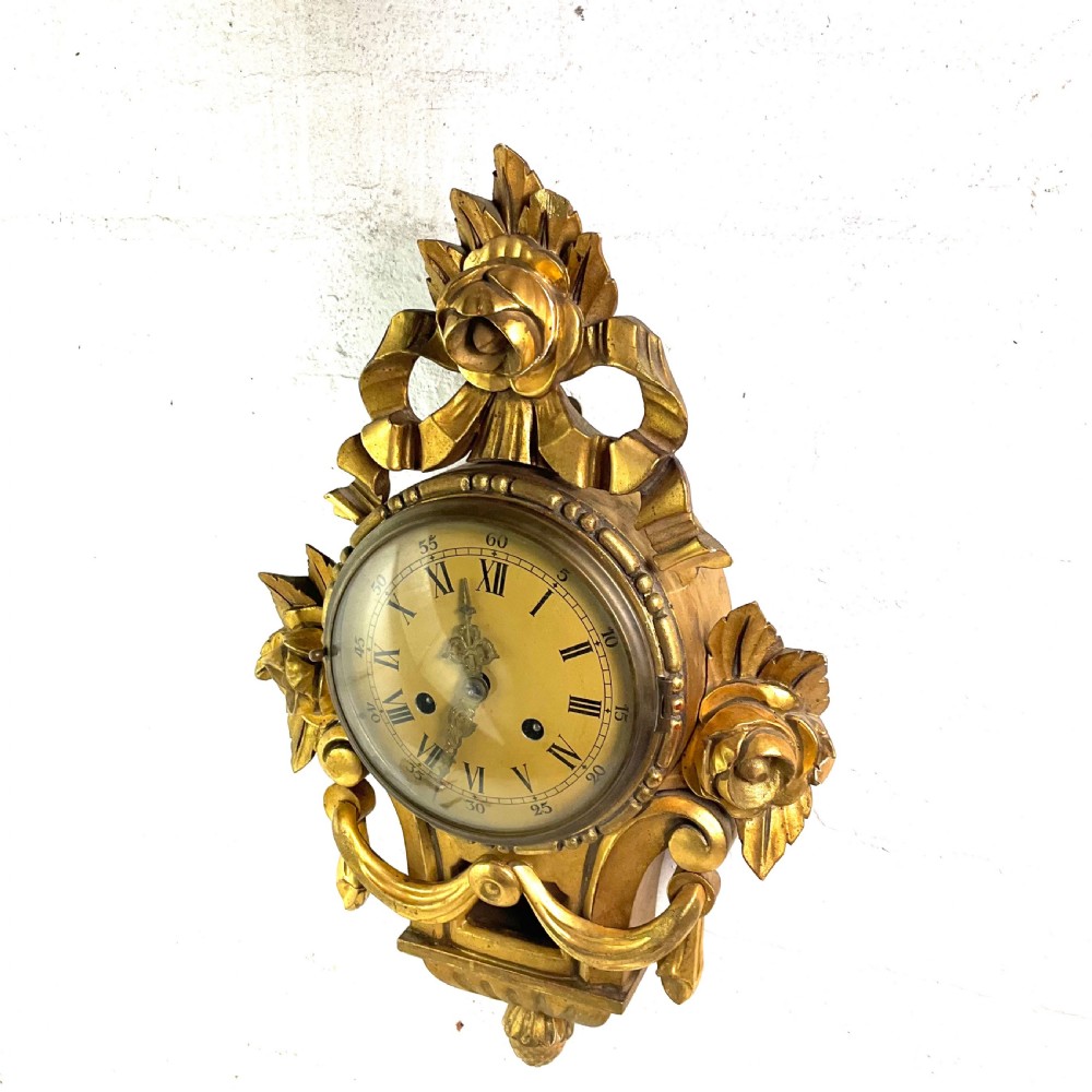 An early 20th-century giltwood cartel wall clock featuring carved floral motifs, a cream dial with Roman numerals, and ornate brass hands. View II