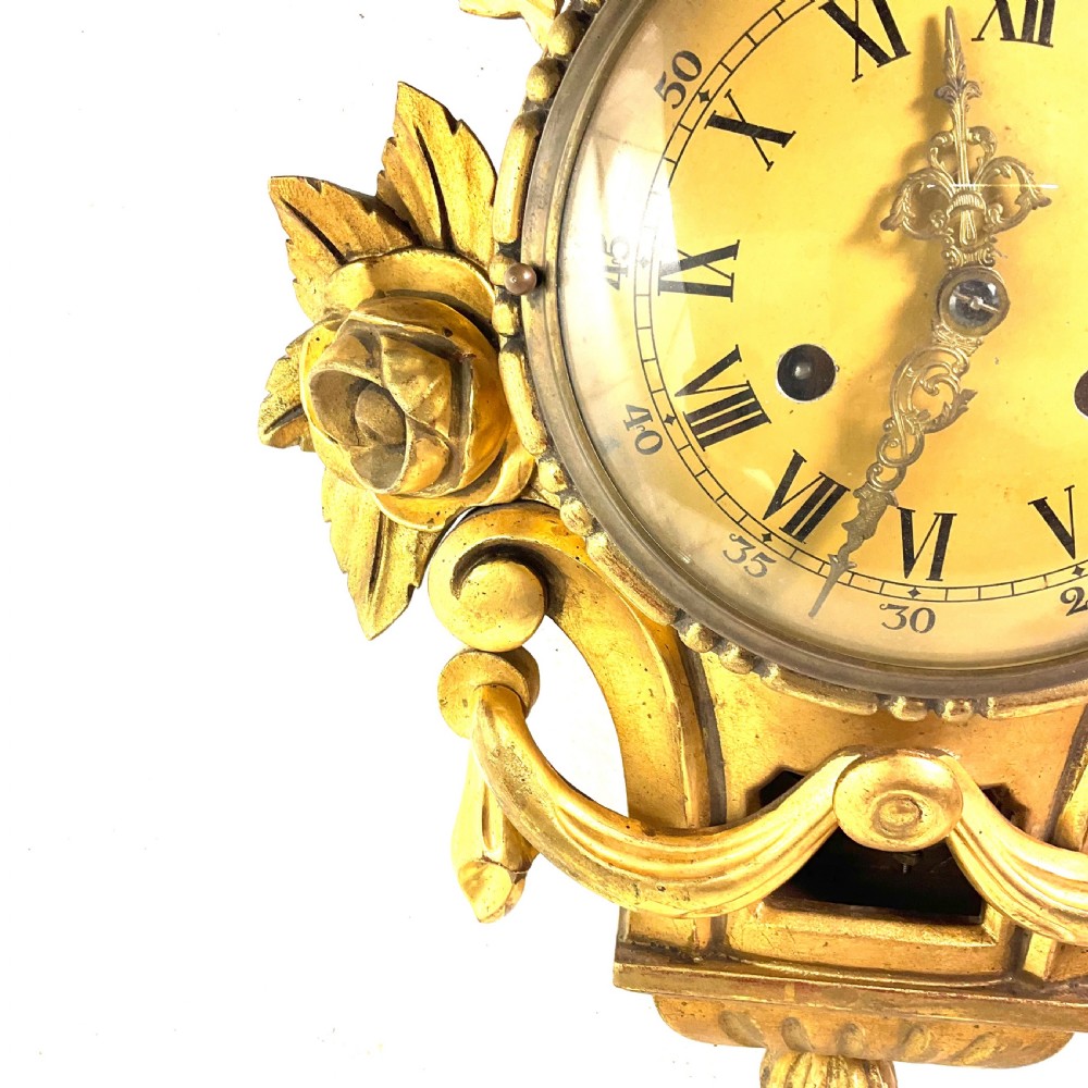 An early 20th-century giltwood cartel wall clock featuring carved floral motifs, a cream dial with Roman numerals, and ornate brass hands. View IV