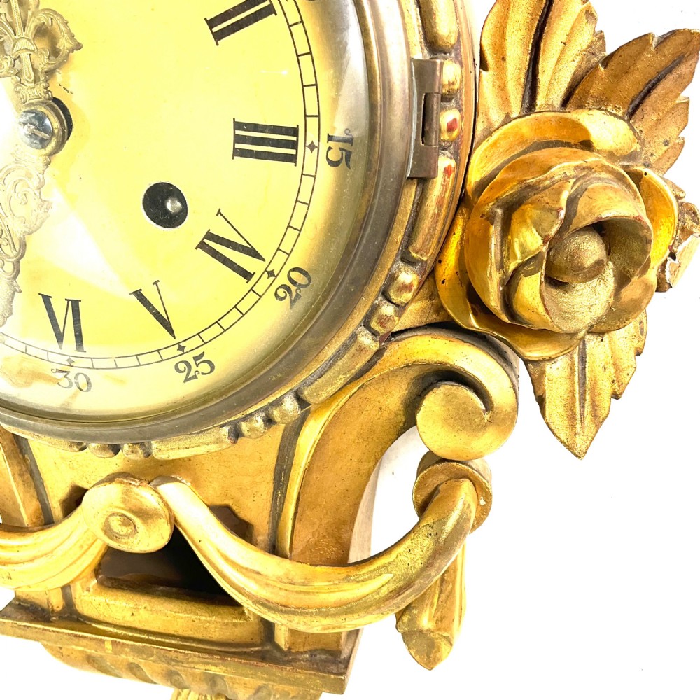 An early 20th-century giltwood cartel wall clock featuring carved floral motifs, a cream dial with Roman numerals, and ornate brass hands.