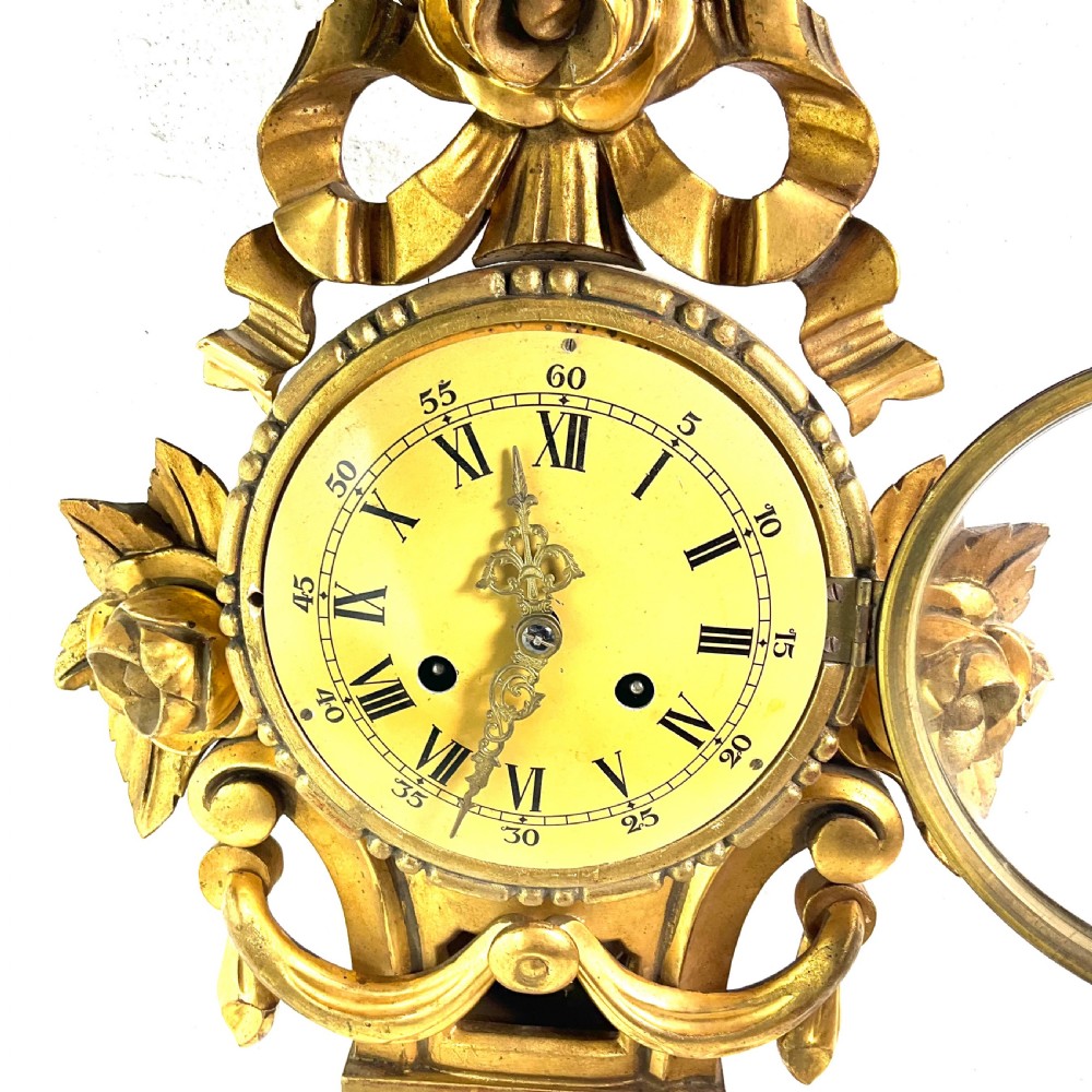 An early 20th-century giltwood cartel wall clock featuring carved floral motifs, a cream dial with Roman numerals, and ornate brass hands. View VI