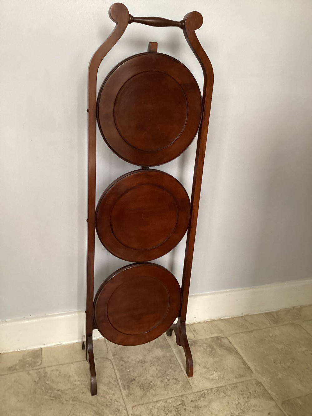 A three-tier Edwardian mahogany folding cake stand featuring circular recessed trays and a curved carrying handle at the top. View VI