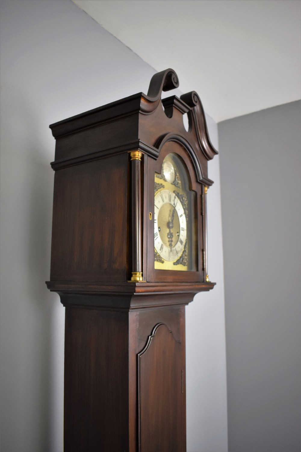 An early 20th-century slim mahogany grandmother clock featuring a swan-neck pediment, brass spandrel dial with Roman numerals, and turned side columns. View IV