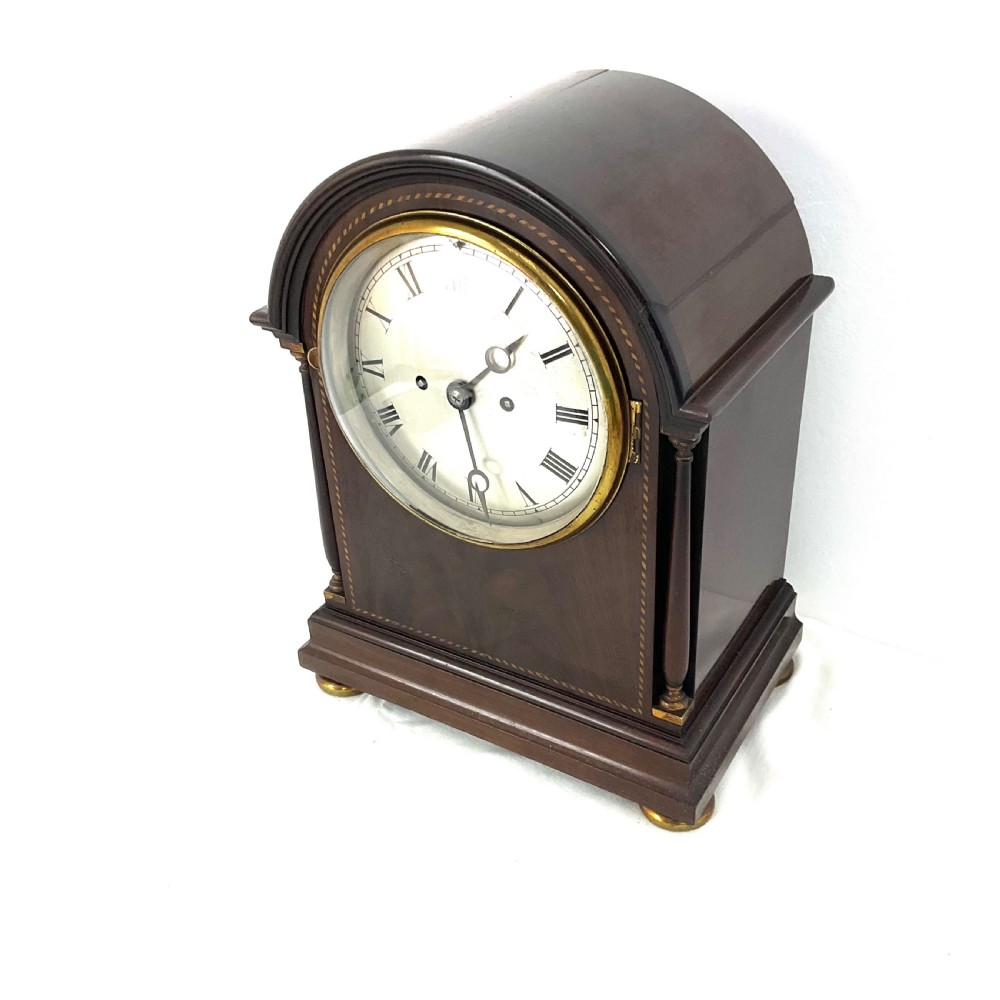 A 19th-century mahogany bracket clock featuring an arched top, brass-framed circular dial with Roman numerals, and brass turned feet. View X