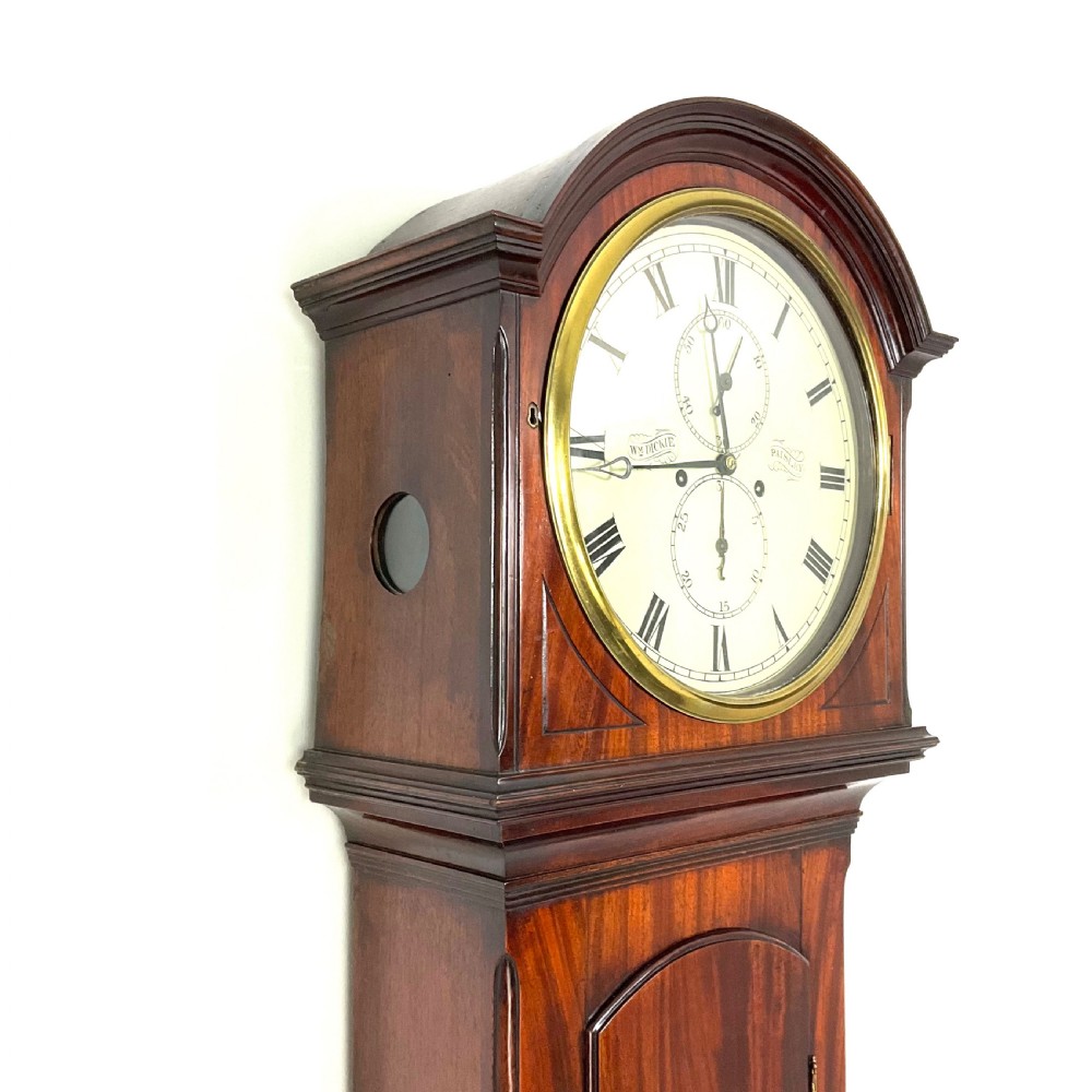 A Regency period flame mahogany longcase clock featuring a circular brass bezel, Roman numerals, subsidiary seconds display, and an arched bonnet. View II