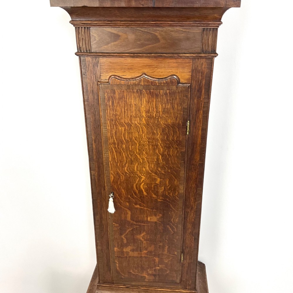 A tall Georgian oak longcase clock featuring a square painted dial, brass column accents, and a panelled wooden case. View VI