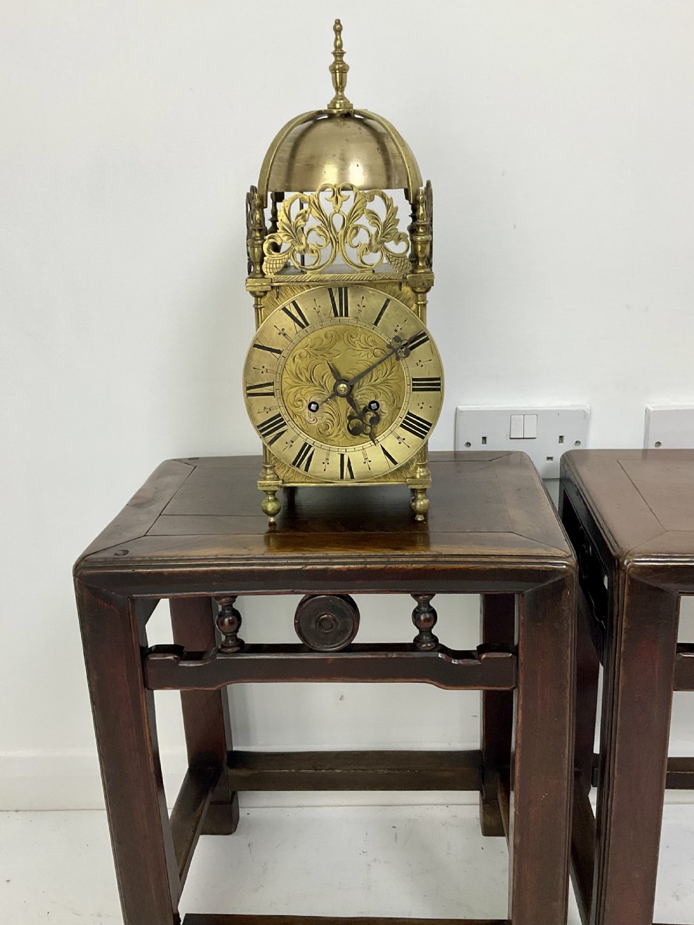 An antique brass lantern clock featuring a top-mounted bell dome, engraved foliate dial with Roman numerals, and ornate pierced frets. View II