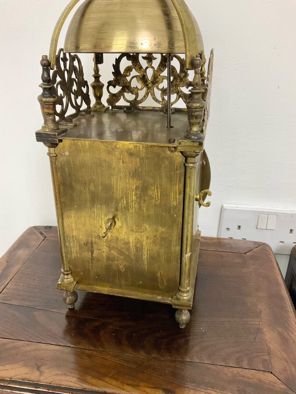 An antique brass lantern clock featuring a top-mounted bell dome, engraved foliate dial with Roman numerals, and ornate pierced frets. View IX