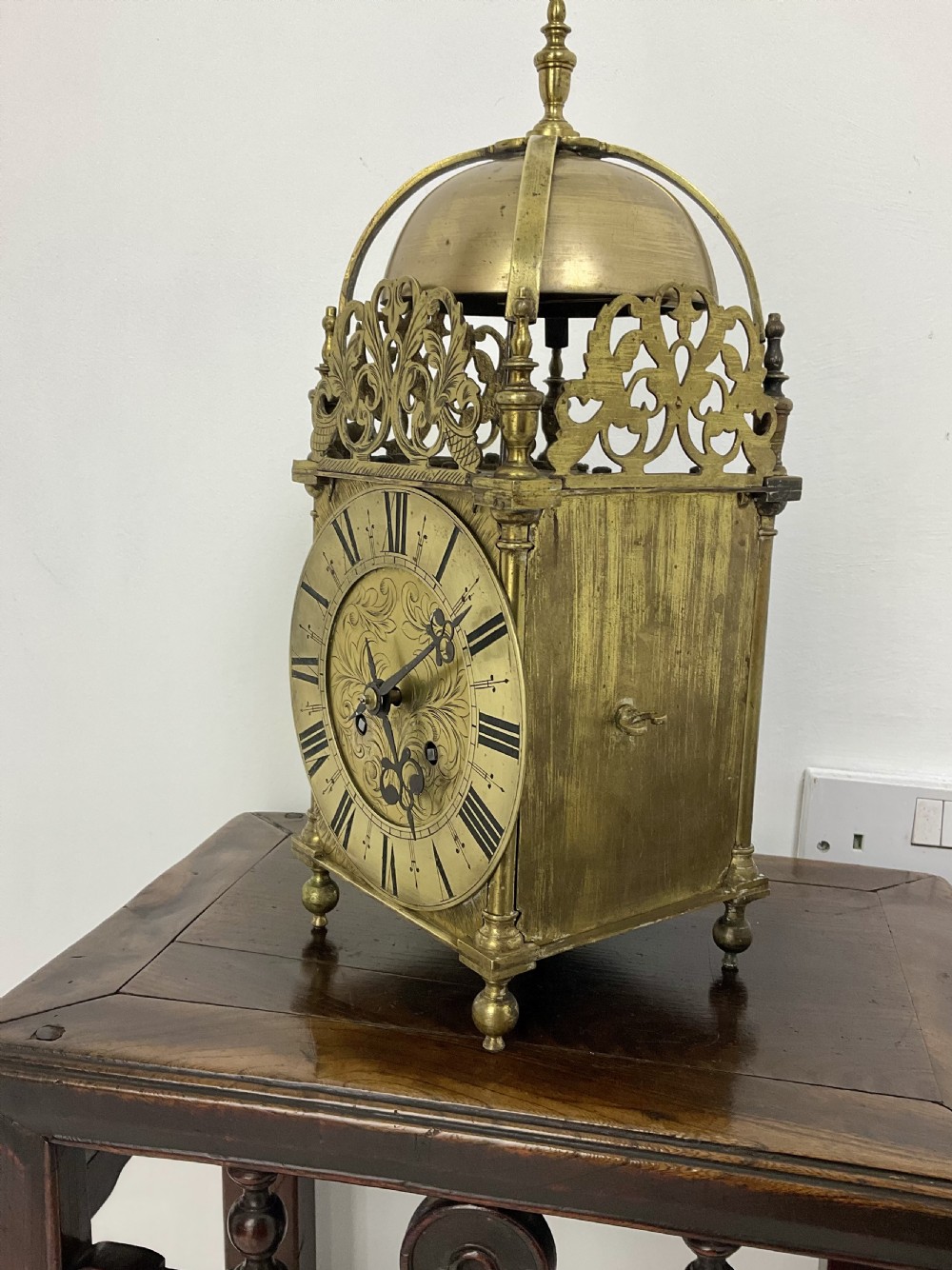 An antique brass lantern clock featuring a top-mounted bell dome, engraved foliate dial with Roman numerals, and ornate pierced frets. View X