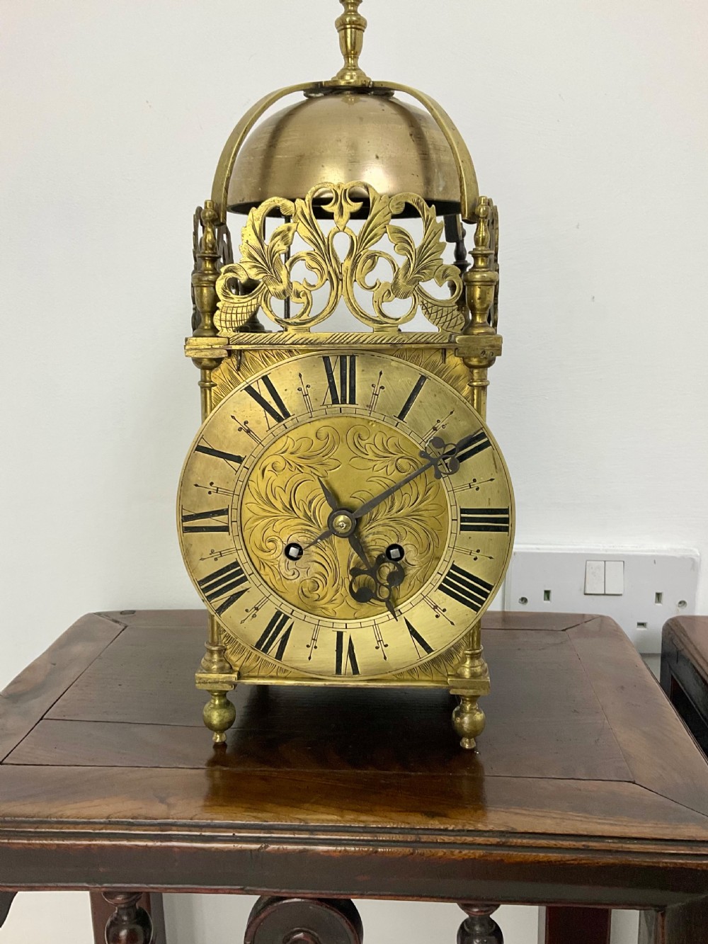 An antique brass lantern clock featuring a top-mounted bell dome, engraved foliate dial with Roman numerals, and ornate pierced frets. View XII
