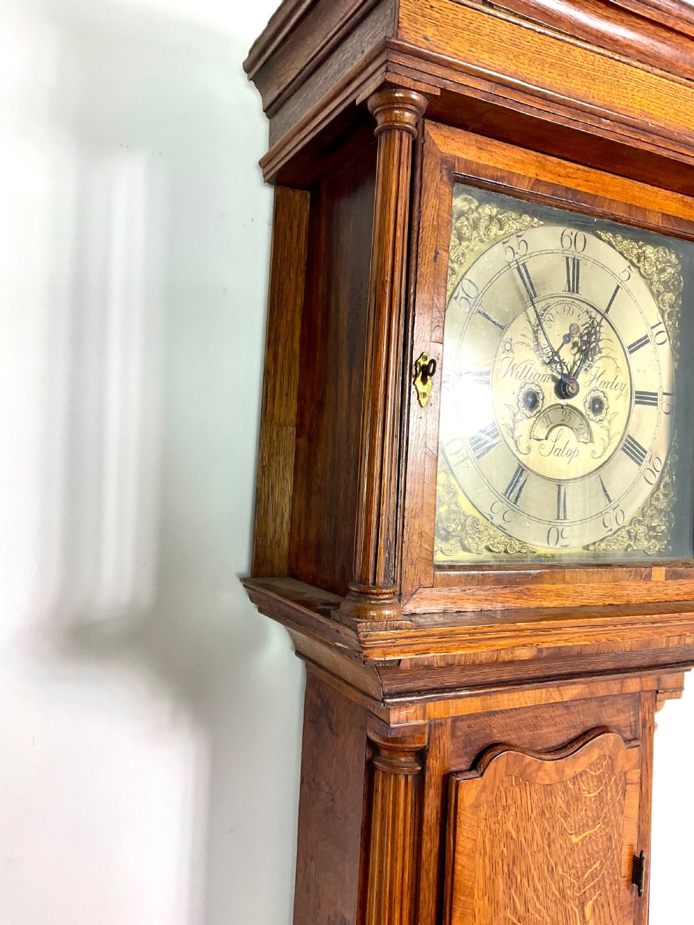 An early Georgian longcase clock featuring a warm oak case with turned hood columns and an engraved brass dial. View IX