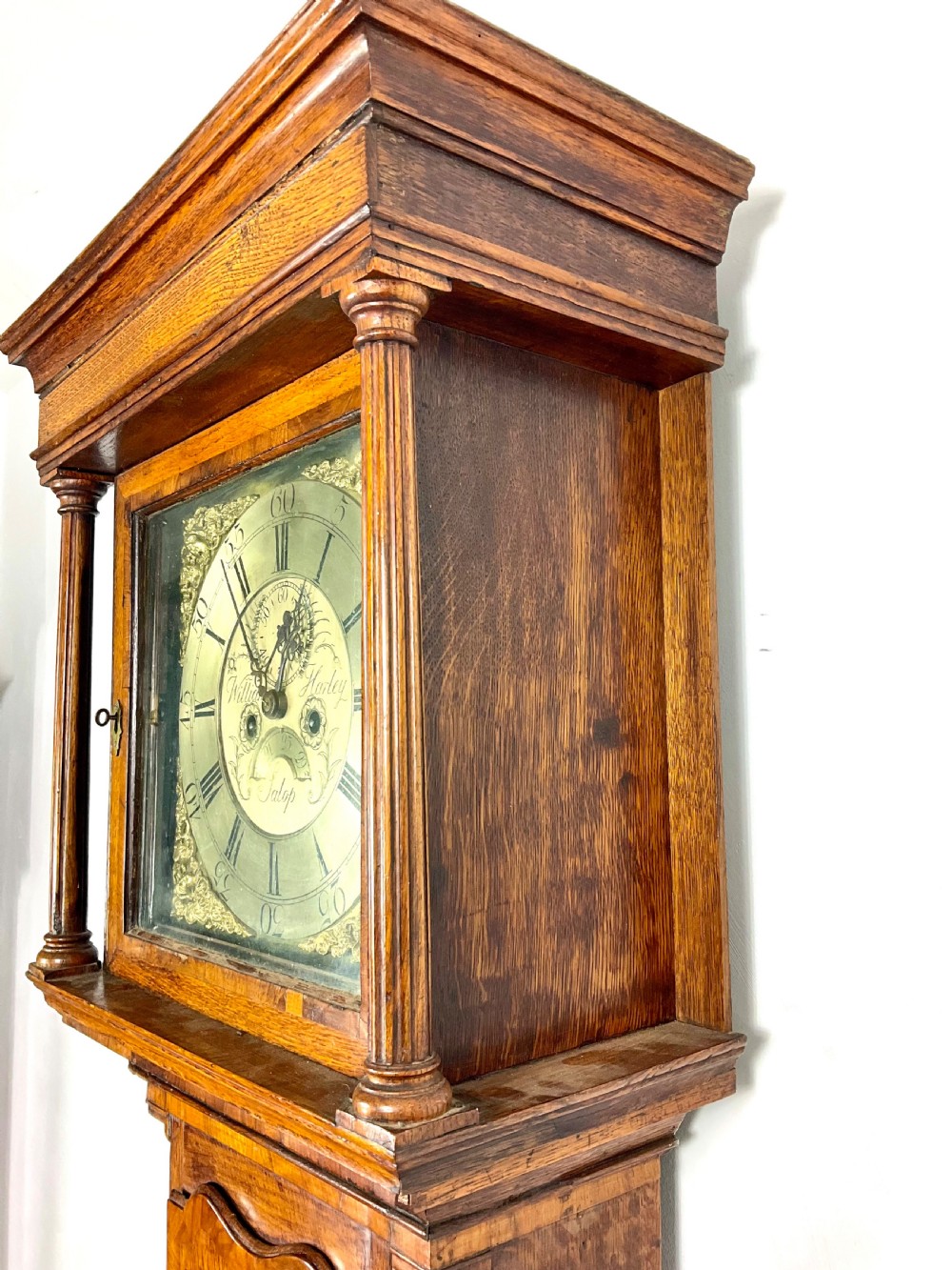 An early Georgian longcase clock featuring a warm oak case with turned hood columns and an engraved brass dial. View X
