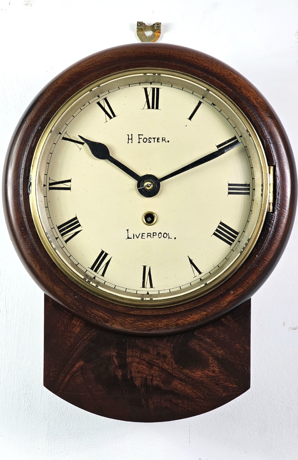 An early Victorian drop dial wall clock featuring a circular off-white dial with Roman numerals, a polished brass bezel, and a rich mahogany casing.