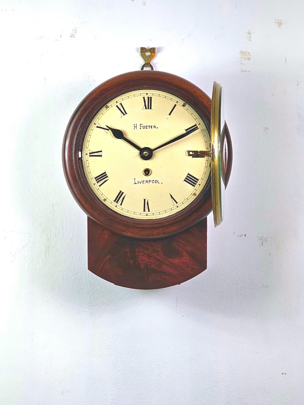 An early Victorian drop dial wall clock featuring a circular off-white dial with Roman numerals, a polished brass bezel, and a rich mahogany casing. View X
