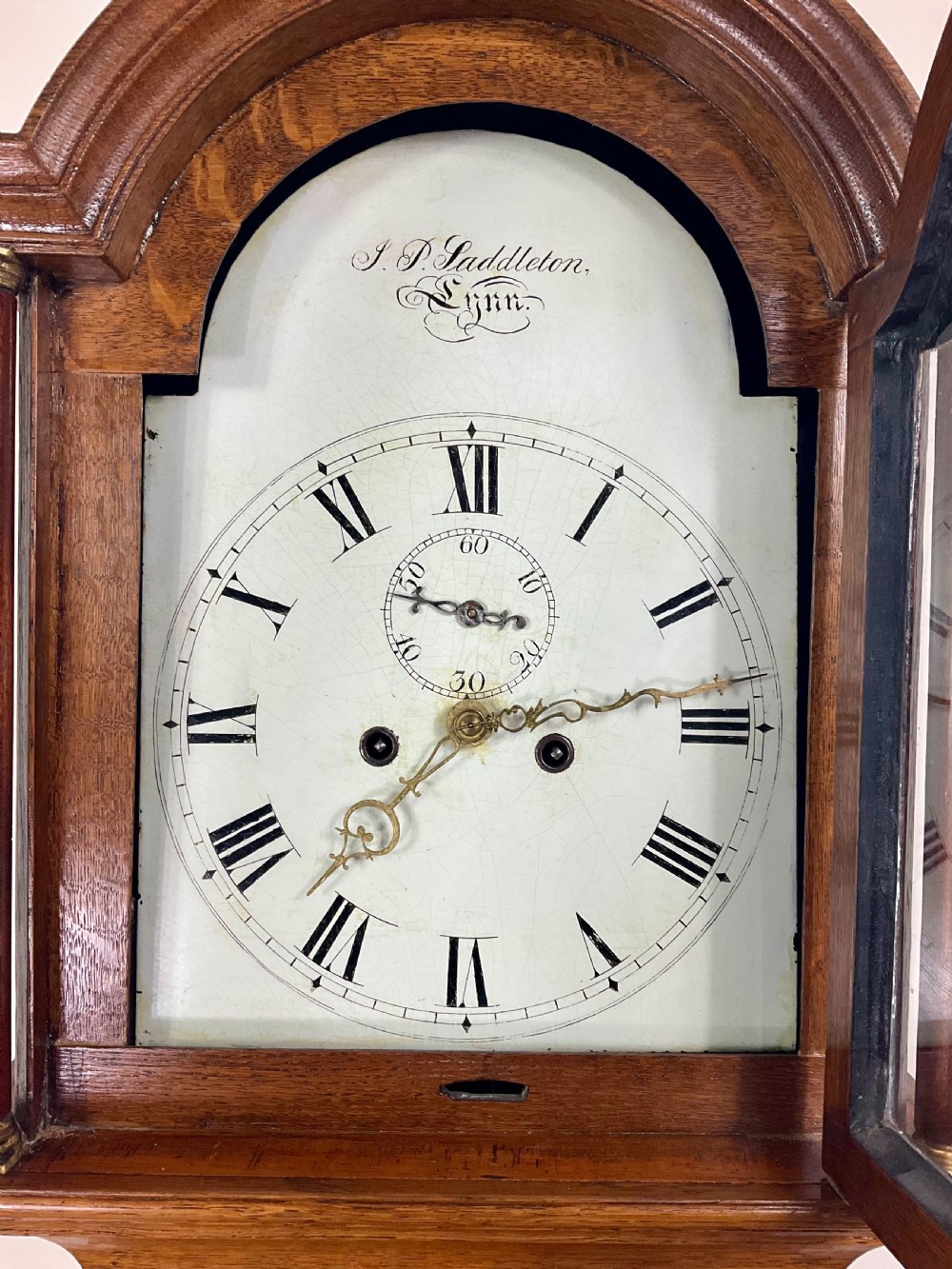 An antique Georgian longcase clock hood crafted from light oak with an arched glass door, brass-capped side columns, and a painted white dial with Roman numerals. View V