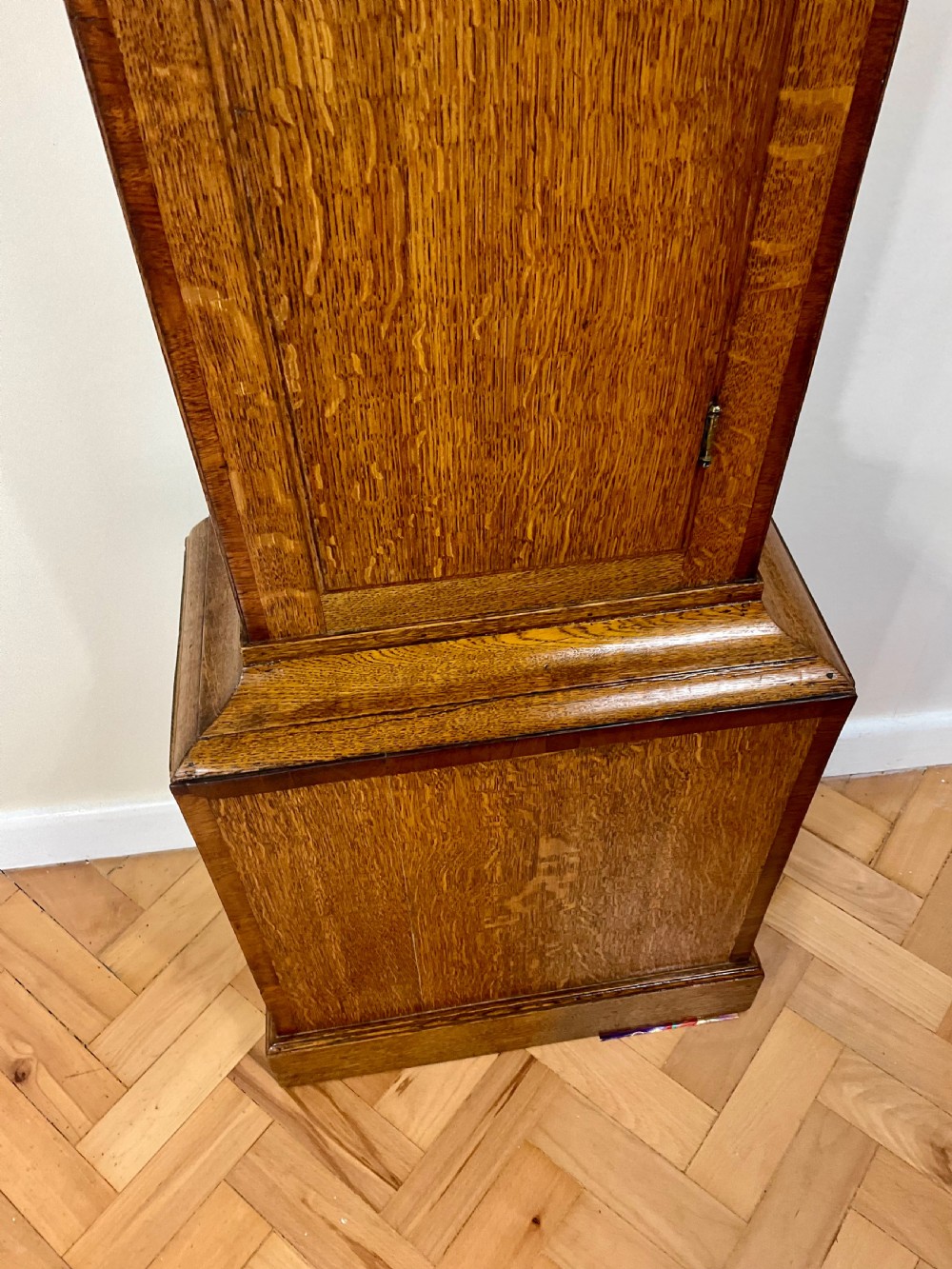 An antique Georgian longcase clock hood crafted from light oak with an arched glass door, brass-capped side columns, and a painted white dial with Roman numerals. View XII